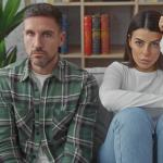 Двойка с напрегнато изражение седи мълчаливо на диван у дома. Мъжът е дистанциран.