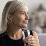 Усмихната жена пие вода от стъклена чаша.